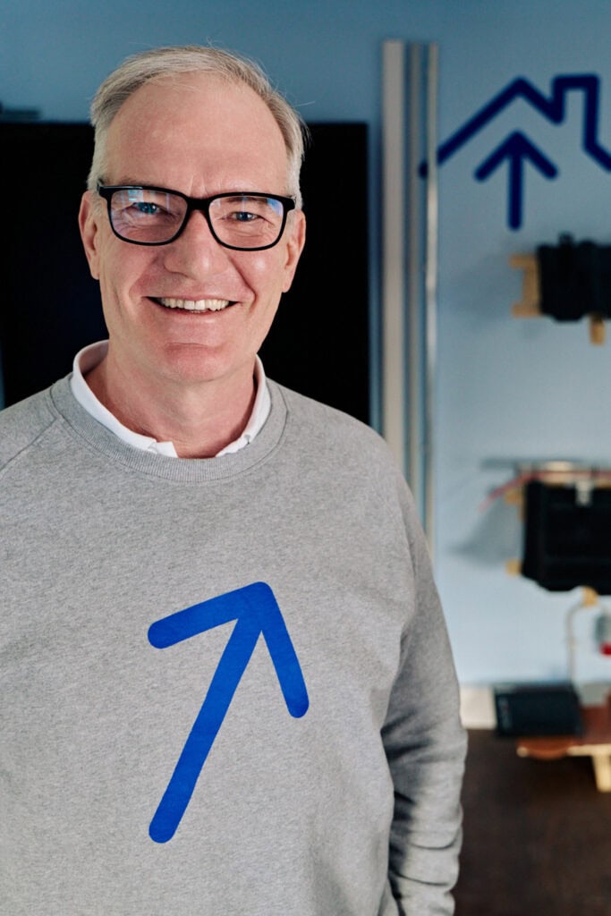 Mann trägt Pullover mit Sonnwärts Logo, freundliches Lächeln, Büro-Umgebung.