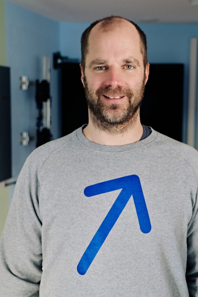 Mann mit grauem Pullover mit blauer Pfeil-Illustration, lächelnd, im Innenraum.