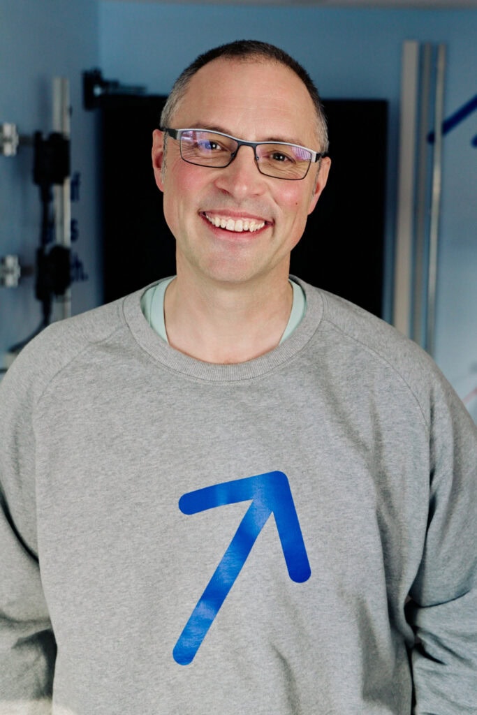 Mann mit Brille und grauem Pullover mit Aufwärtspfeil, lächelnd.