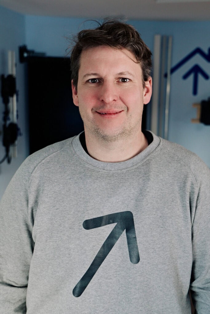 Mann trägt grauen Pullover mit Sonnwärts-Logo, lächelnd im Büro.