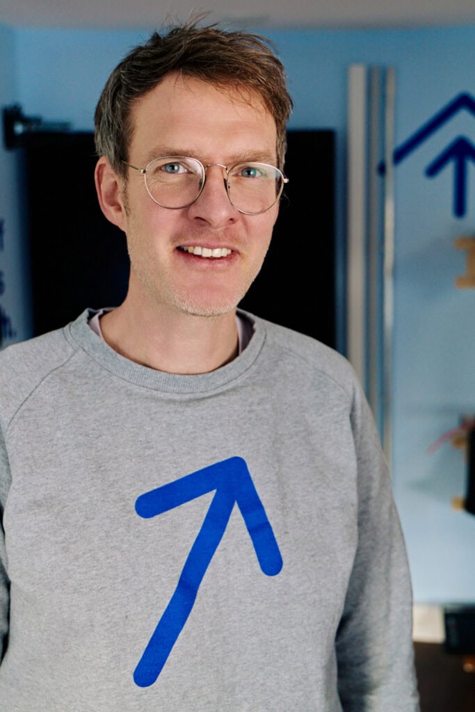 Freundlicher Mann mit Brille und grauem Pullover vor Sonnwärts-Logo.